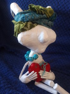 Paper and tape puppet with horns and a headwrap, a string of skull beads and holding a fabric heart.